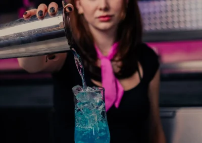 A bartender pouring a bright blue liquid over ice from a cocktail shaker into a tall glass, with a pink background