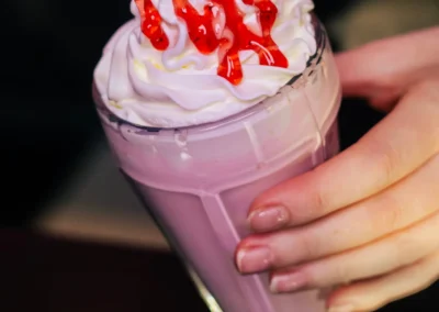 A hand drizzling red strawberry syrup over whipped cream on a pink milkshake