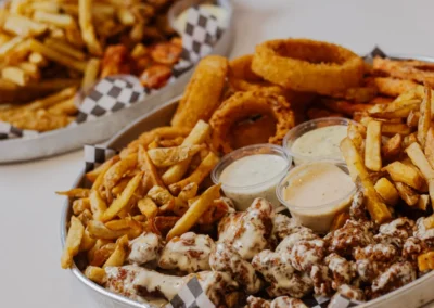 A large platter featuring a variety of fried foods including french fries, onion rings, and pieces of chicken drizzled with sauce, alongside dipping cups