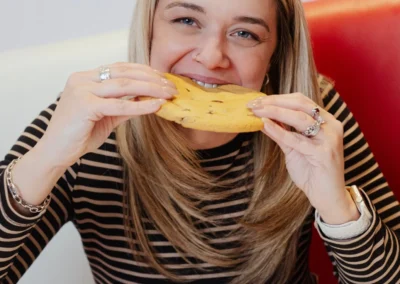 A woman in a striped shirt smiling while taking a bite out of a large chocolate chip cookie in a diner booth